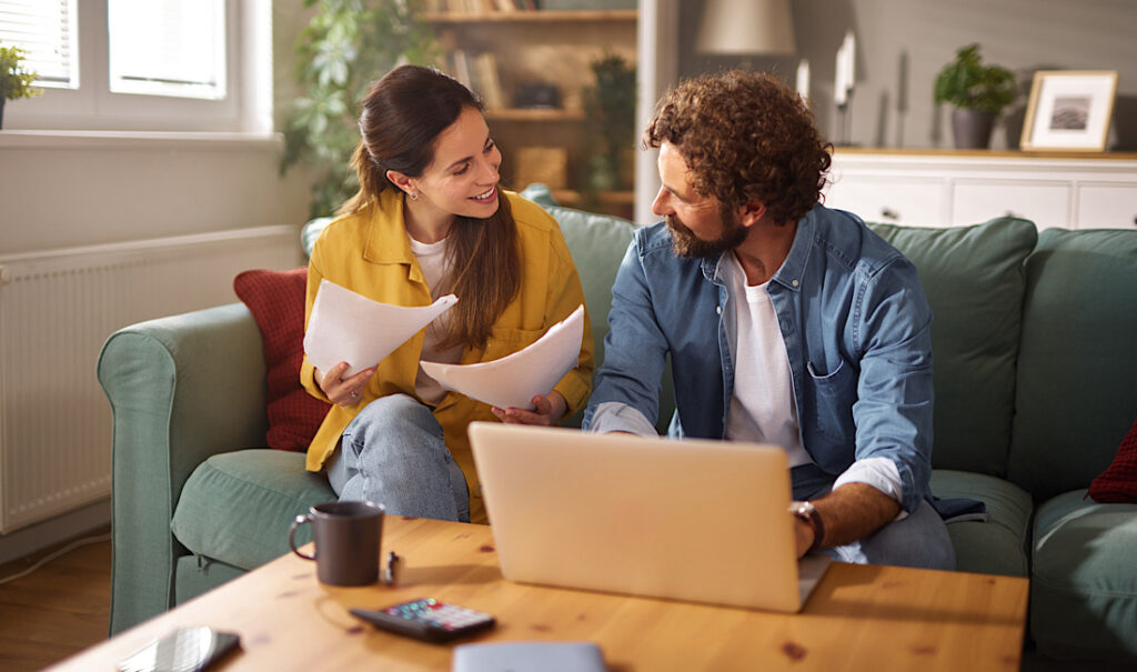 A couple sits together on their couch discussing various life insurance plans. They review important documents while a laptop is open on the coffee table, creating a warm atmosphere in Castle Rock, CO.